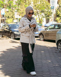 Woman in a beige coat and hijab standing on a sidewalk with cars and trees in the background