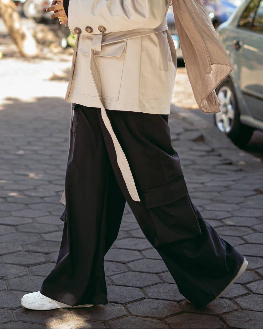 Woman walking outdoors wearing a beige trench coat, black pants, and sunglasses.