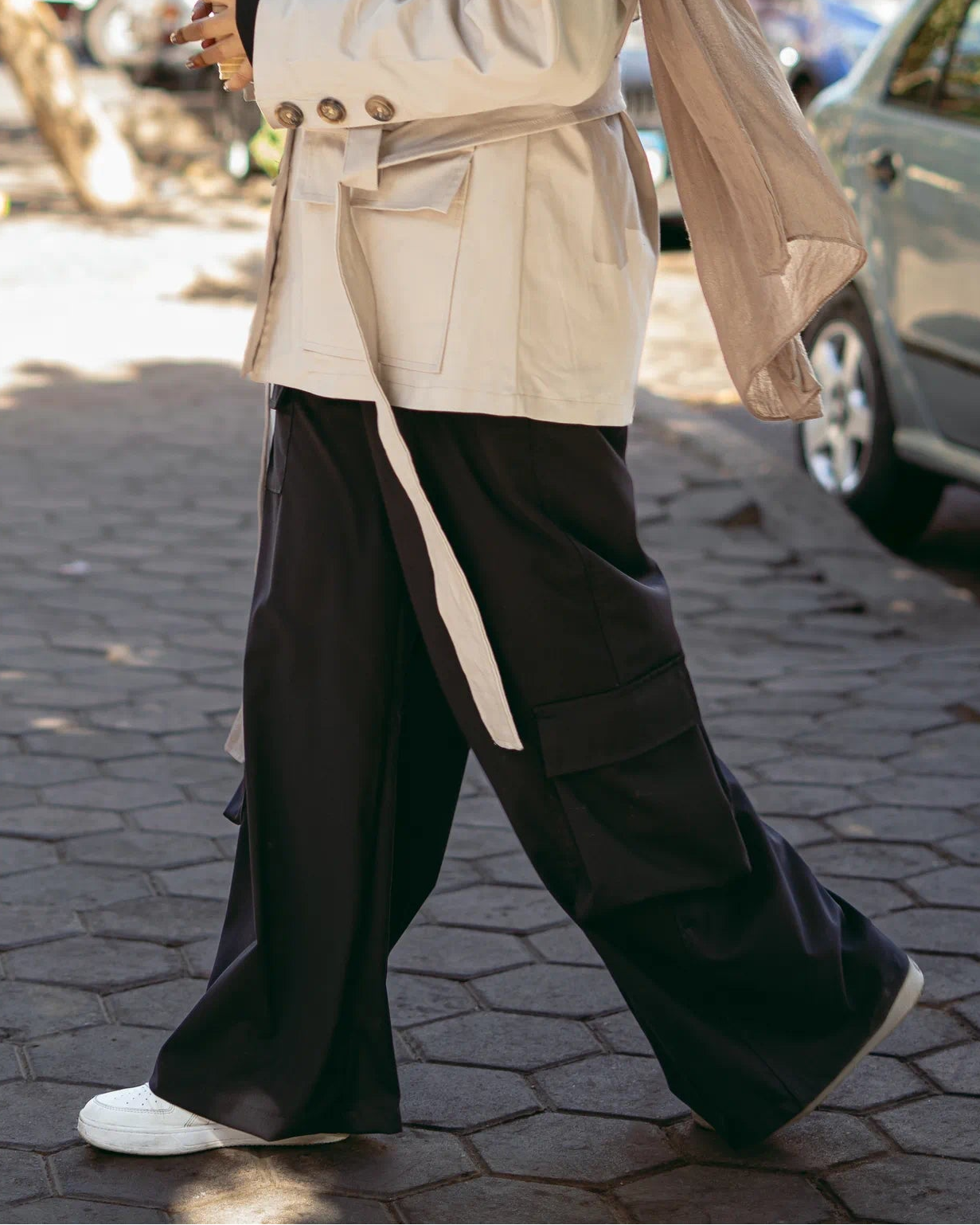 Woman walking outdoors wearing a beige trench coat, black pants, and sunglasses.