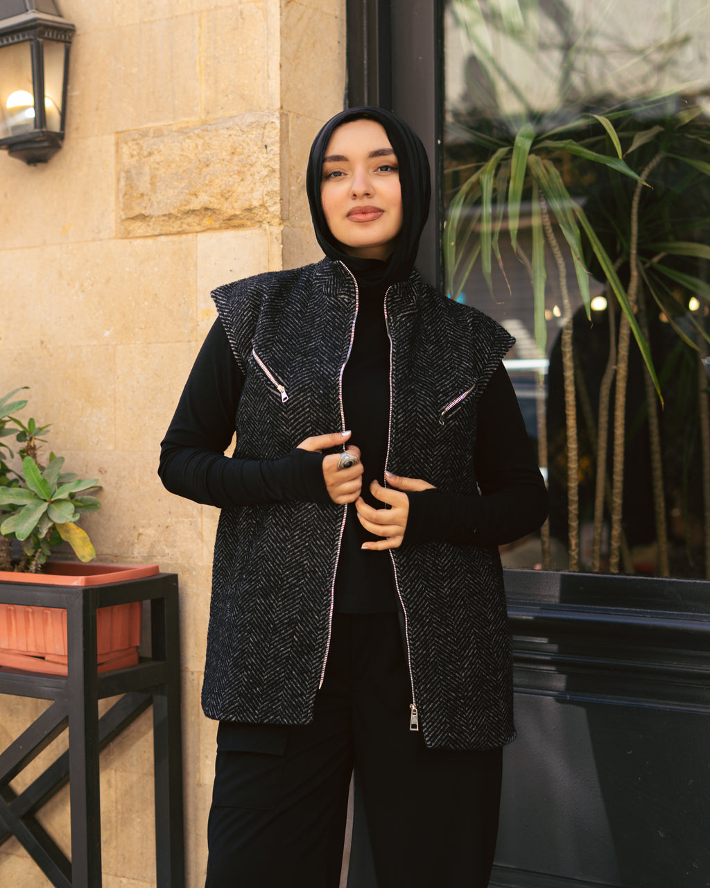 Woman wearing a black vest over a black outfit with a hood, standing outdoors.