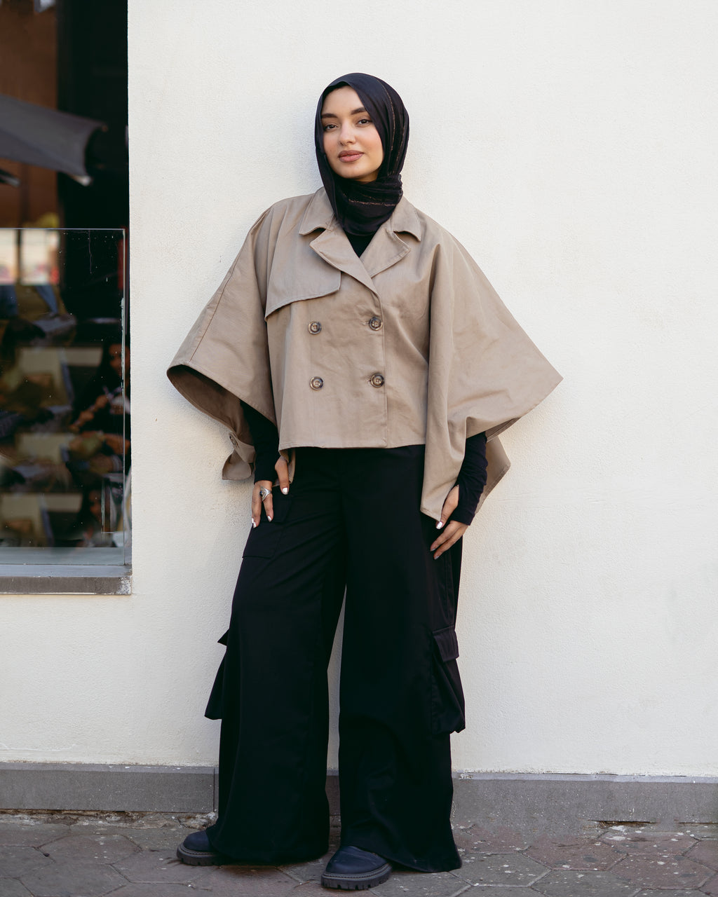 Person wearing a beige cape over a black outfit against a white wall.