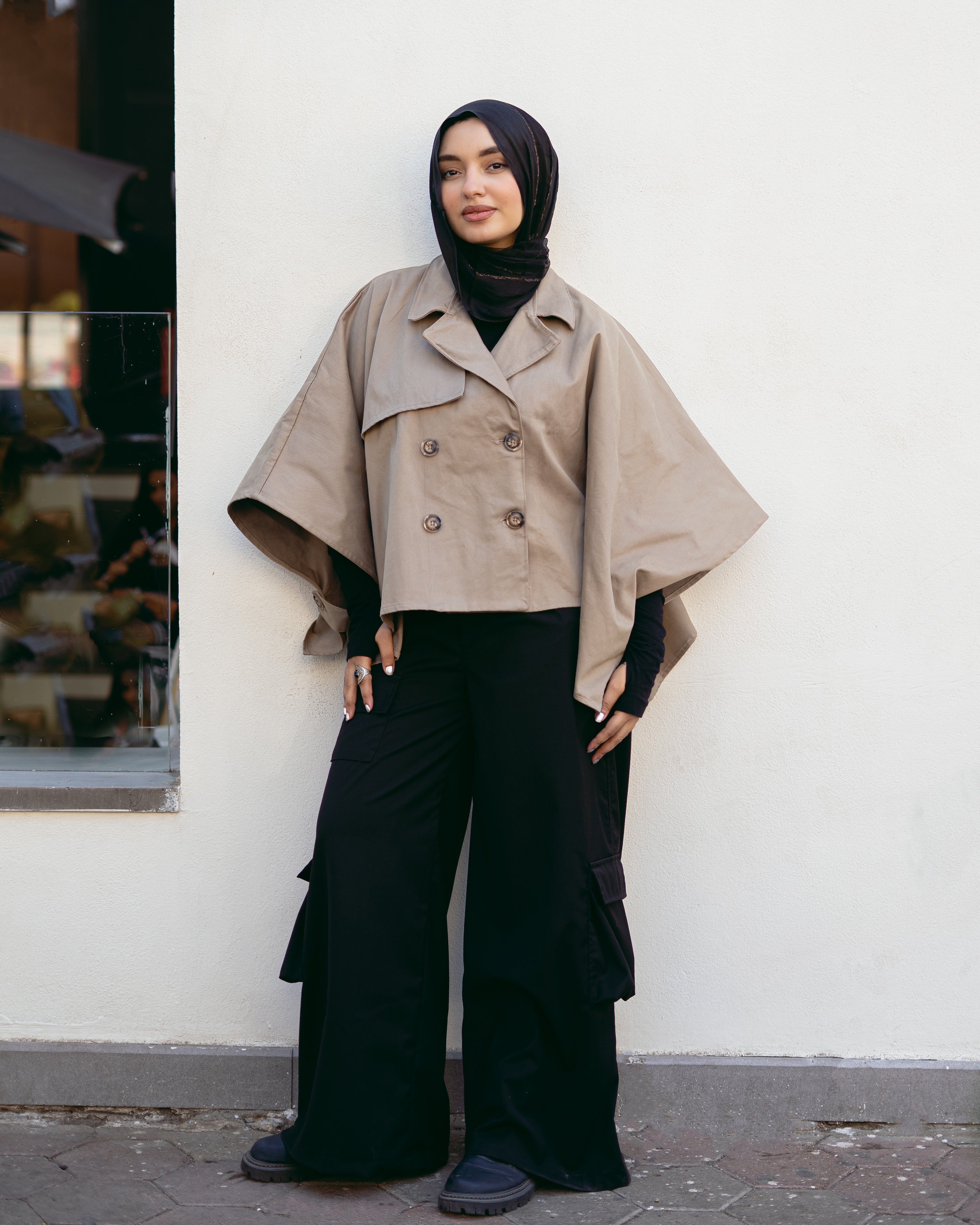 Person wearing a beige cape over a black outfit against a white wall.