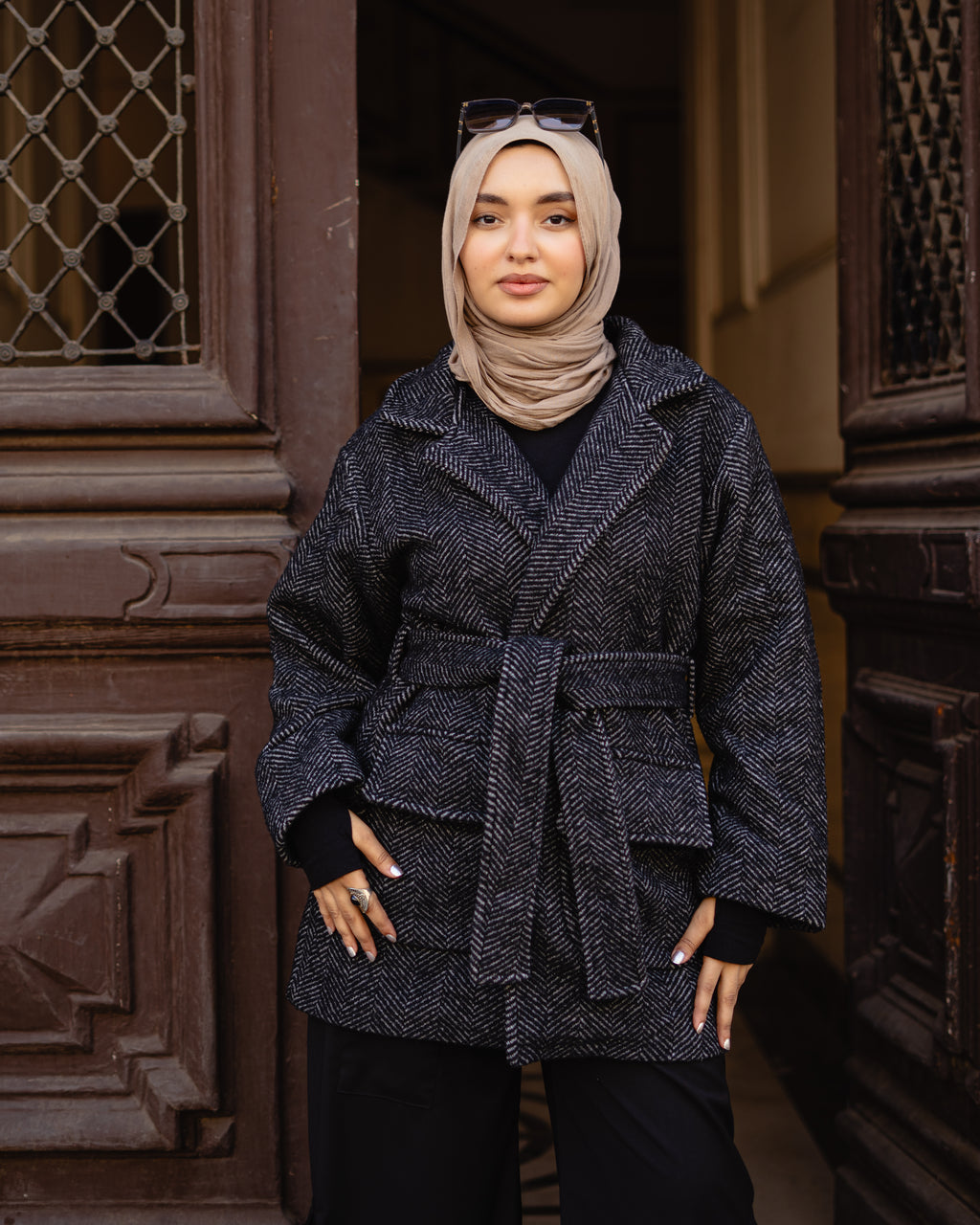 Woman wearing a dark blue textured coat and beige hijab standing in front of a decorative door.