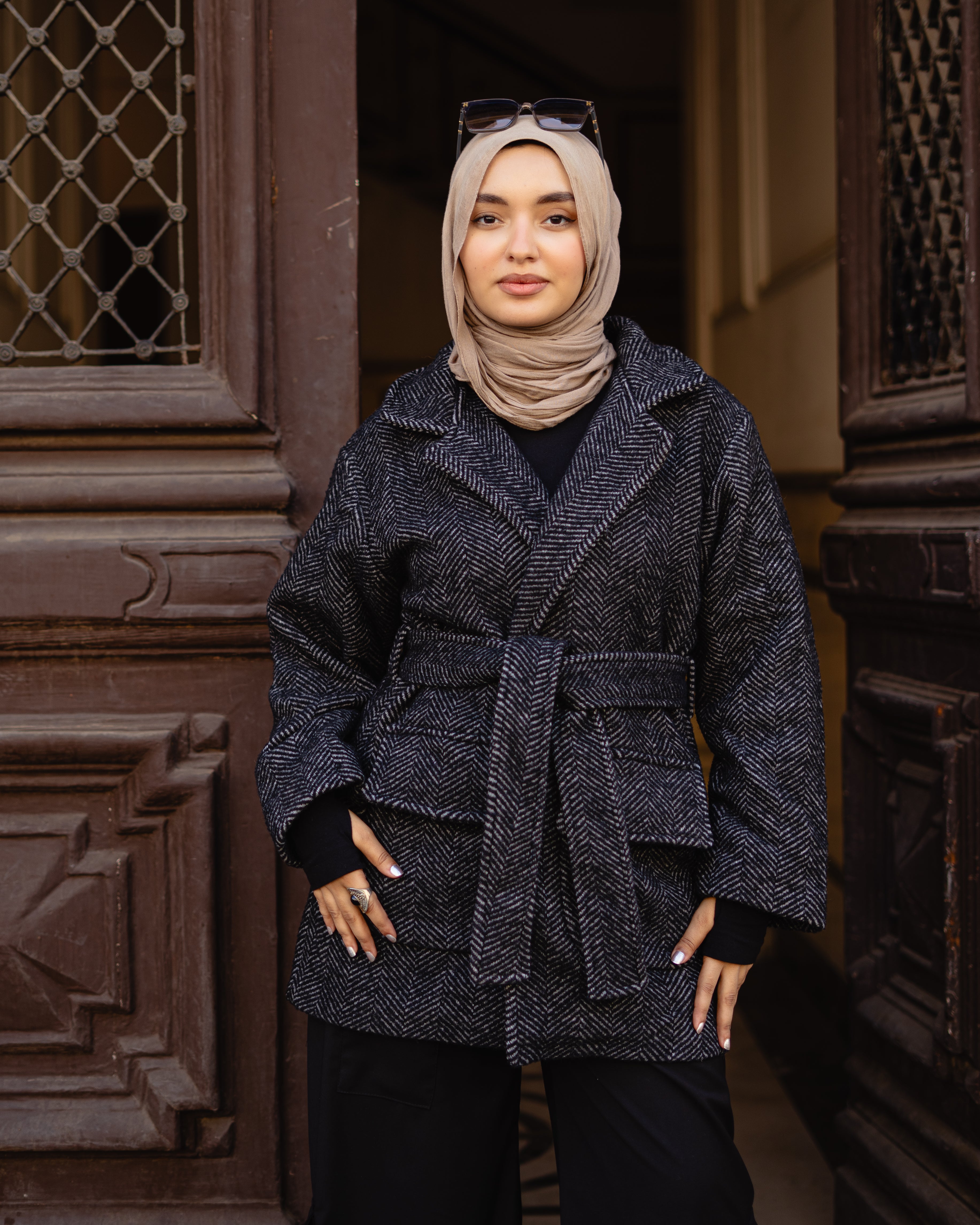 Woman wearing a dark blue textured coat and beige hijab standing in front of a decorative door.
