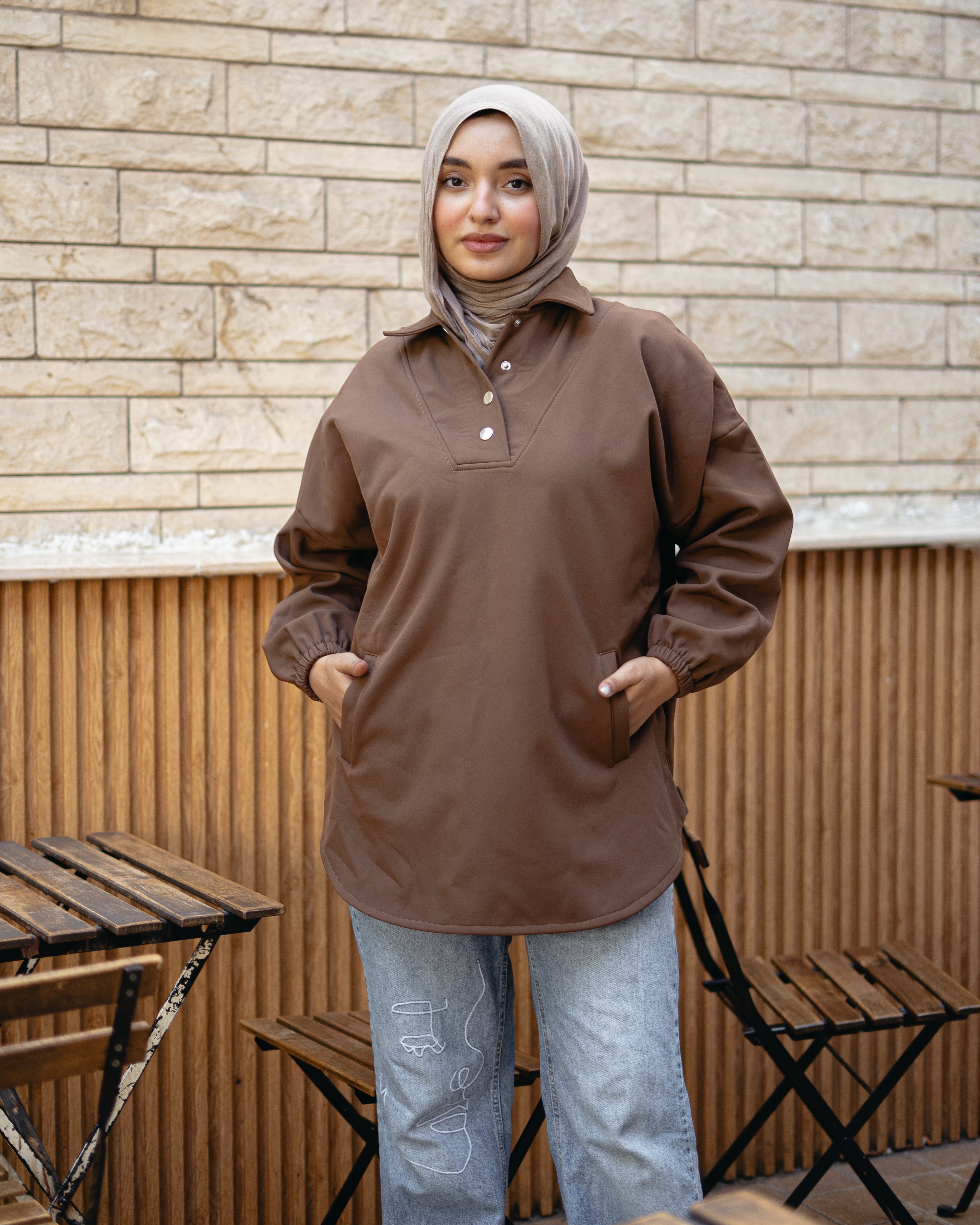 Woman wearing a brown jacket and hijab standing outdoors with a stone wall background