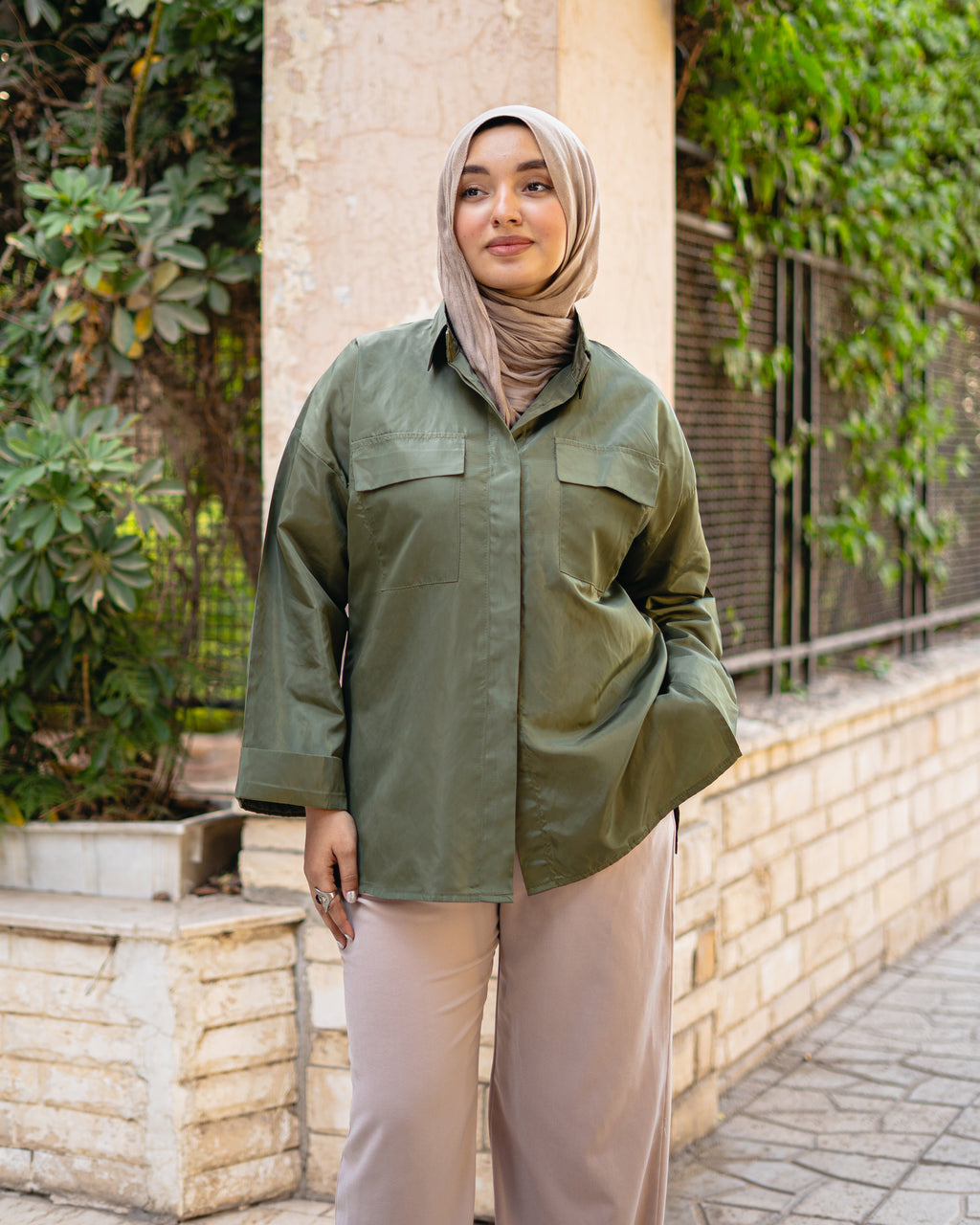 Woman wearing an olive green jacket and beige pants standing outdoors with plants in the background