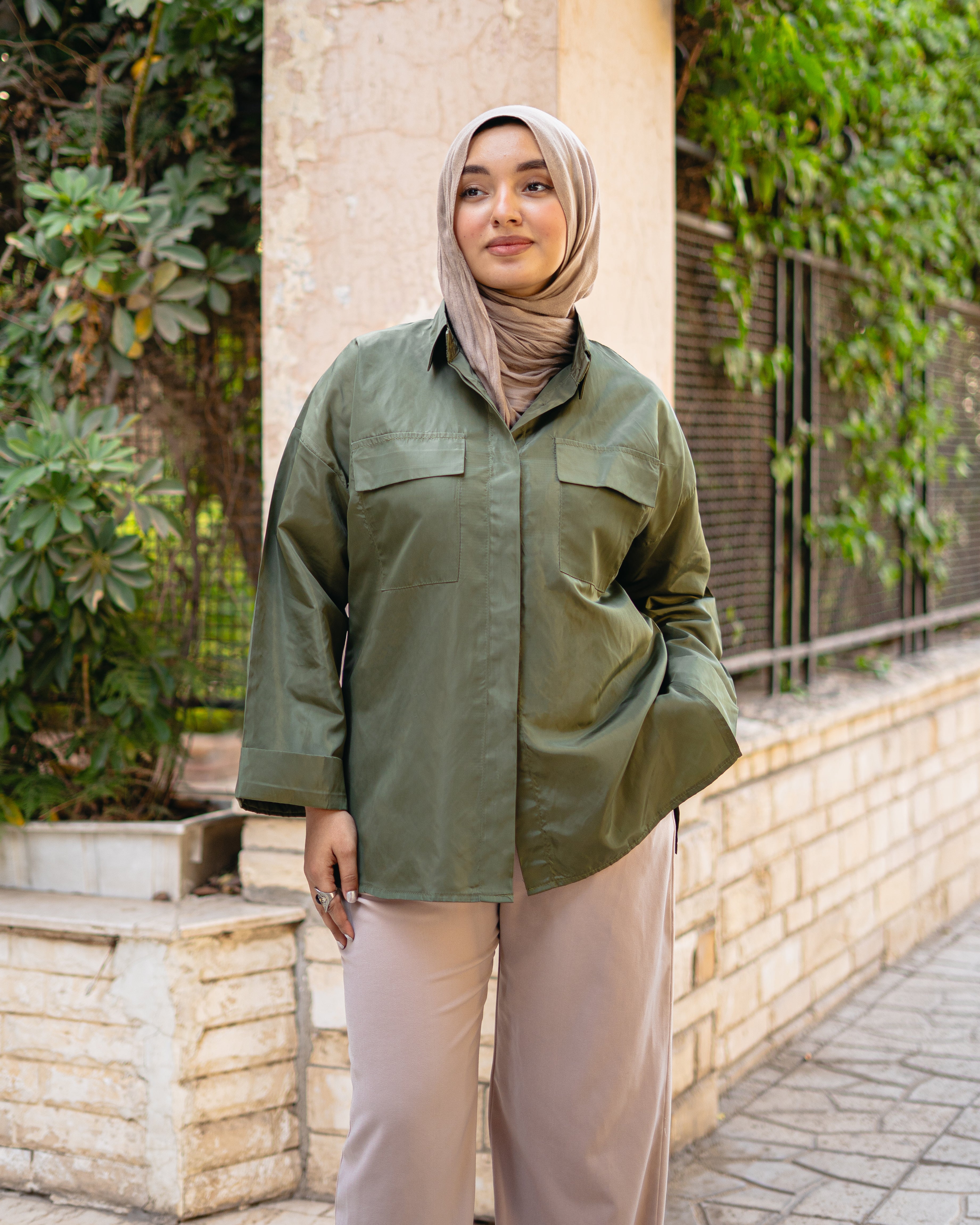 Woman wearing an olive green jacket and beige pants standing outdoors with plants in the background